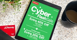 An iPad displaying the JCPenney Cyber Monday 2022 advertisement on a counter with a JCPenney card, a cup of coffee, and a plant