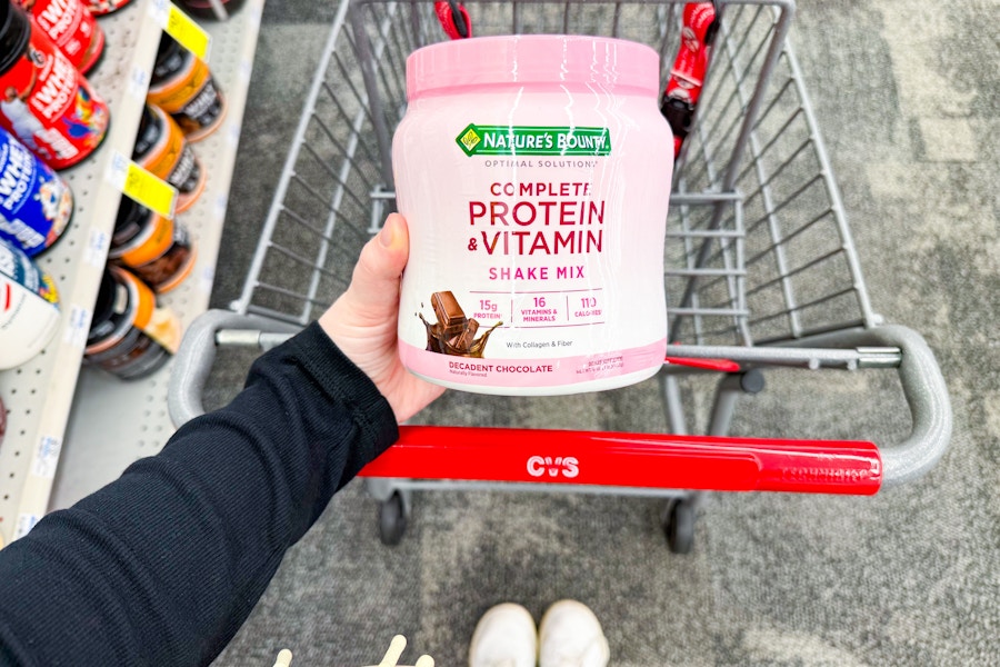 person holding a tub of protein powder over a cvs cart
