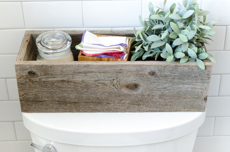 Wood box on the back of the toilet filled with a candle, indoor house plant, and a box of cloth diapers.