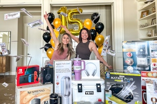 The Krazy Coupon Lady's co founders, Heather and Joanie tossing money over a pile of items they are giving away for KCL's 15th birthday