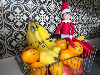 an elf on the shelf doll holding sharpie with faces marked on bananas and oranges in a basket of fruit 