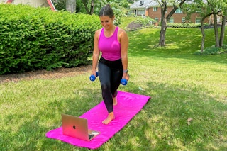A woman stands on a pink yoga mat and does exercises from a laptop