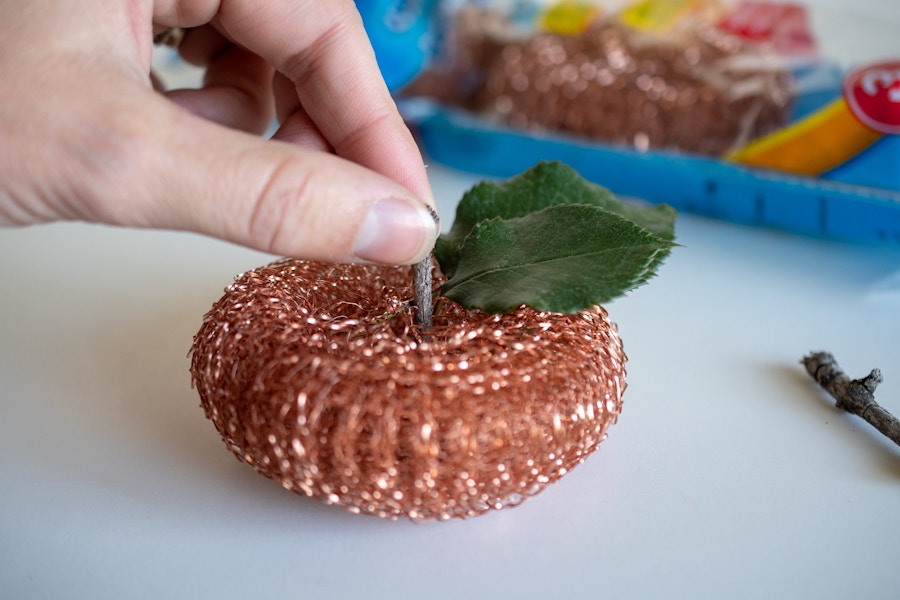 A person putting a leaf and a twig in a kitchen scrubber, making it look like a pumpkin.