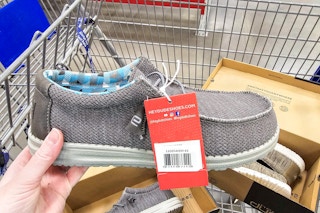 person holding a grey hey dude shoe over a cart with 2 boxes of hey dude shoes