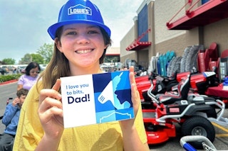 a young girl wearing a blue plastic Lowe's hat and holding up a sign that says "I love you to bits, Dad!" outside of a Lowe's store