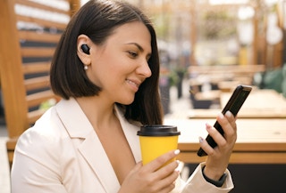A person wearing earbuds looking at a phone and holding a cup of coffee