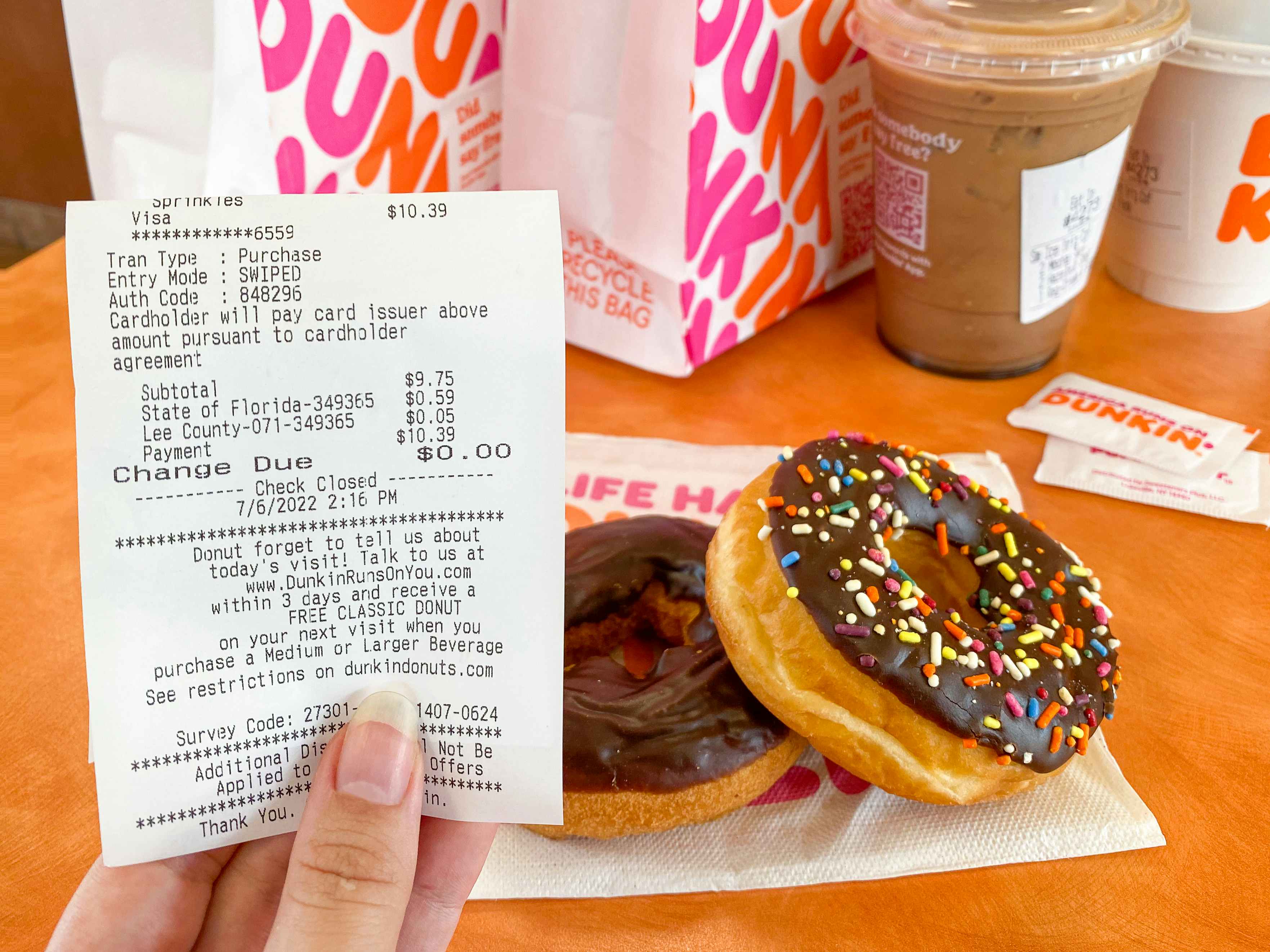 A person's hand holding a receipt offering a free donut for taking the survey printed on the bottom with some donuts, two Dunkin Donuts c...