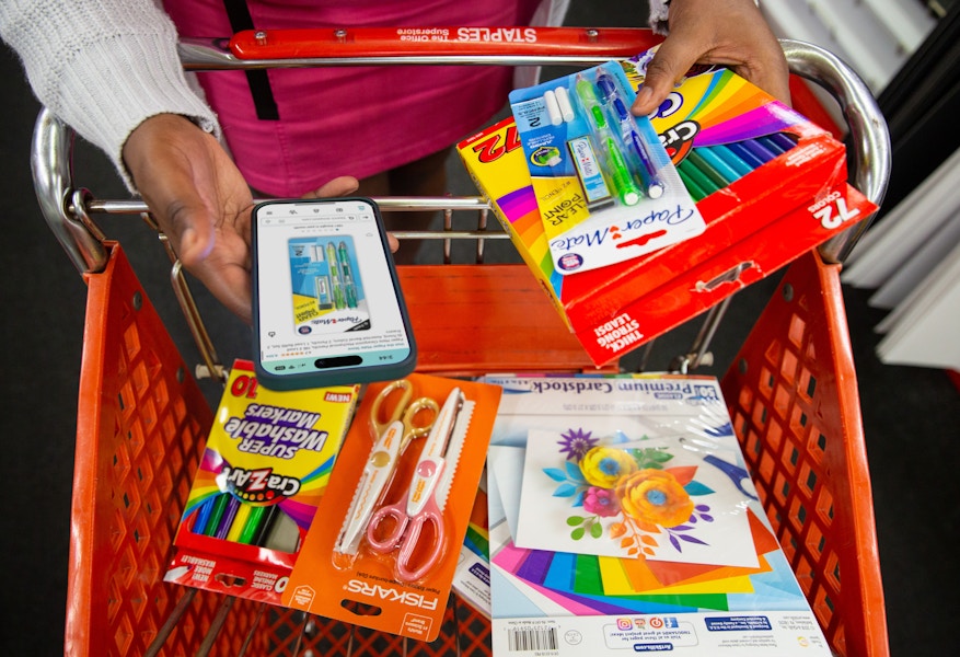 someone holding a phone looking up prices on amazon for some school supplies in a Staples cart