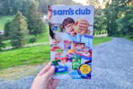 person holding the sams club instant savings book in front of trees