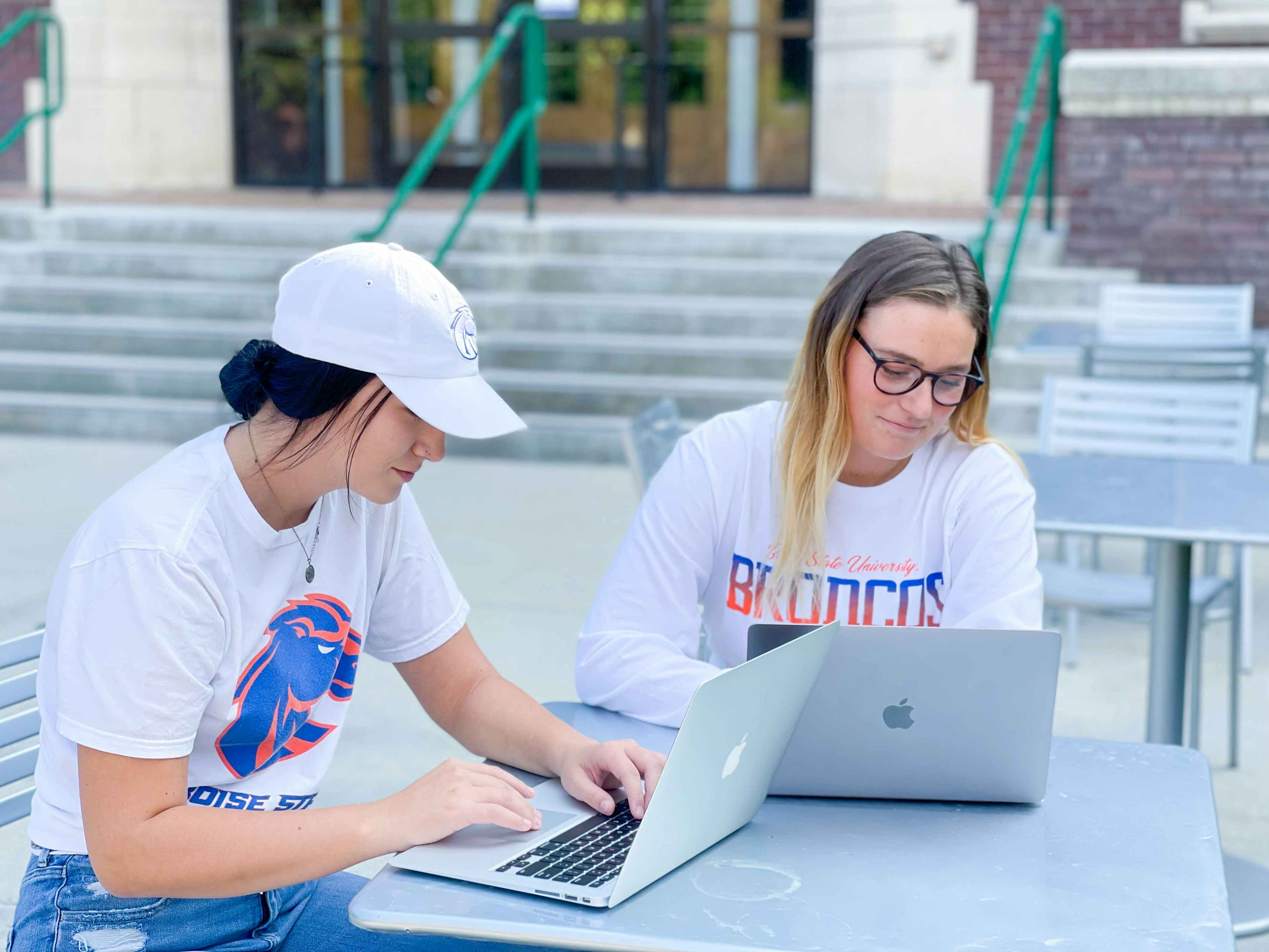 Two college students sitting outside at a table, working on their laptops.