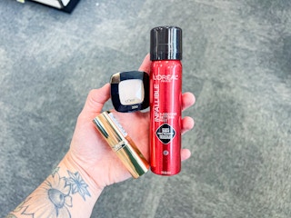 person holding loreal lipstick, eyeshadow, and setting spray