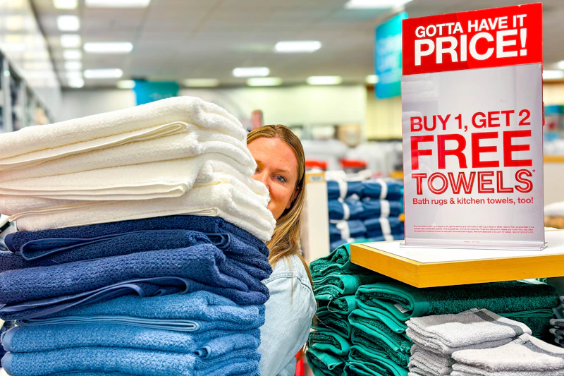 A person holding up a stack of towels next to a buy 1 get 2 free towels sale sign inside JcPenney. 