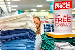 A person holding up a stack of towels next to a buy 1 get 2 free towels sale sign inside JcPenney.