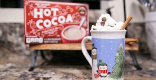 A christmas mug with hot cocoa, whip cream, chocolate and a cinnamon stick. Behind it is a box of hot cocoa mix