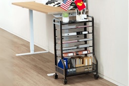 A rolling metal 7-tier storage cart stands next to a standing desk with teacher stuff on it