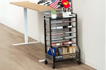 A rolling metal 7-tier storage cart stands next to a standing desk with teacher stuff on it