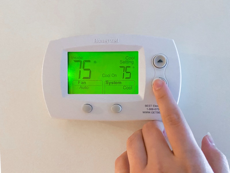 A person's hand going to turn the temperature down on the thermostat which shows 75 degrees Fahrenheit.