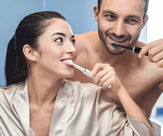 A woman and man brushing their teeth with an electric toothbrush.