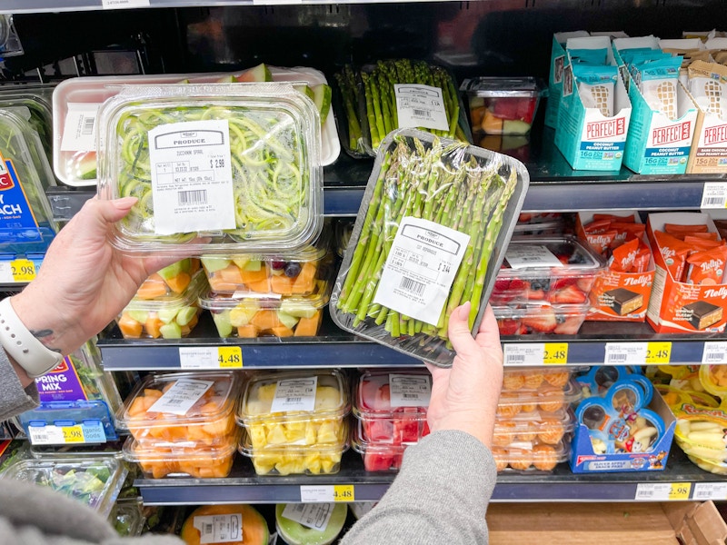 A person's hands holding up prepackaged zucchini spirals and asparagus in front of the prepackaged produce shelf in a grocery store.