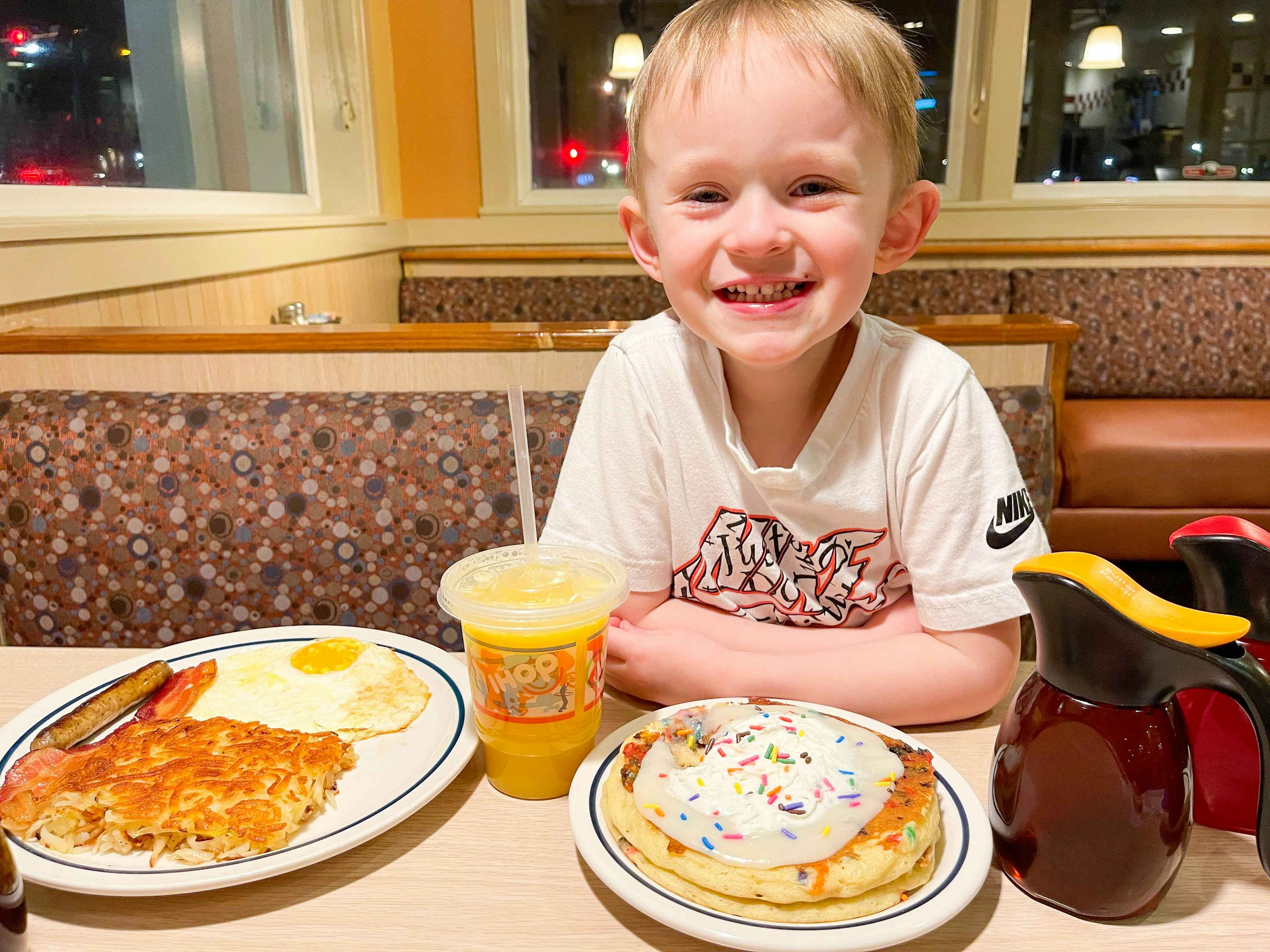 A child smiling and resting their elbows on the table of a booth at iHop. There is a plate of eggs, sausage, and hash browns, a plate of ...