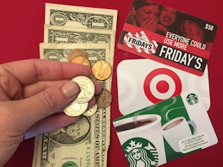 A person holding three dollar bills and change in their hand above a stack of gift cards.