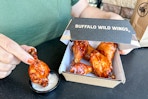 A person holding a Buffalo Wild Wing chicken wing with other chicken wings in a to go box.