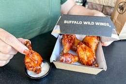 A person holding a Buffalo Wild Wing chicken wing with other chicken wings in a to go box.