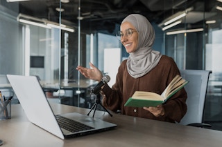 A person sitting at a desk, reading a book into a microphone to make a recording for an audiobook site