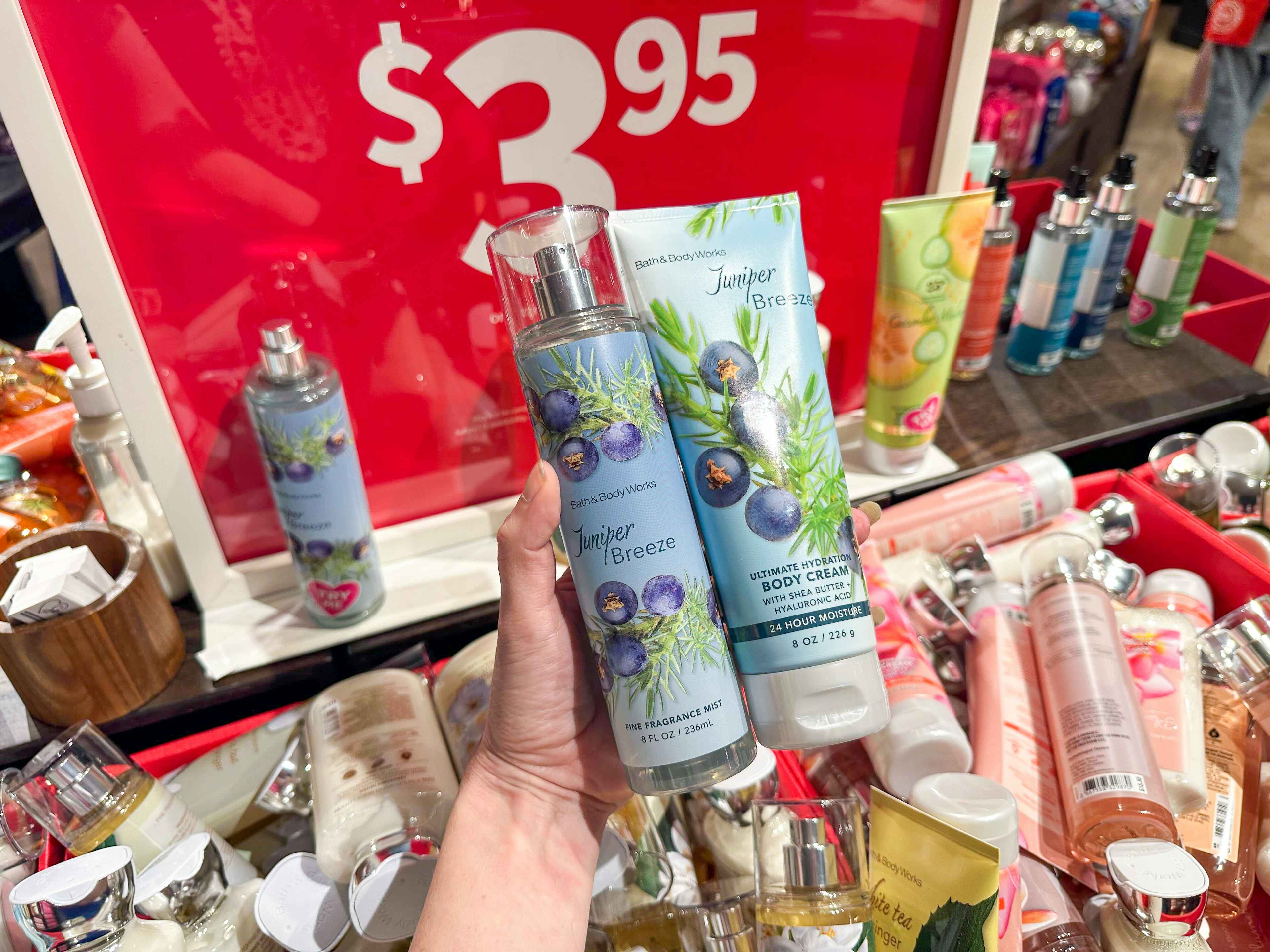 A bath and body works spray and lotion held out by hand in front of a sale sign and other products.
