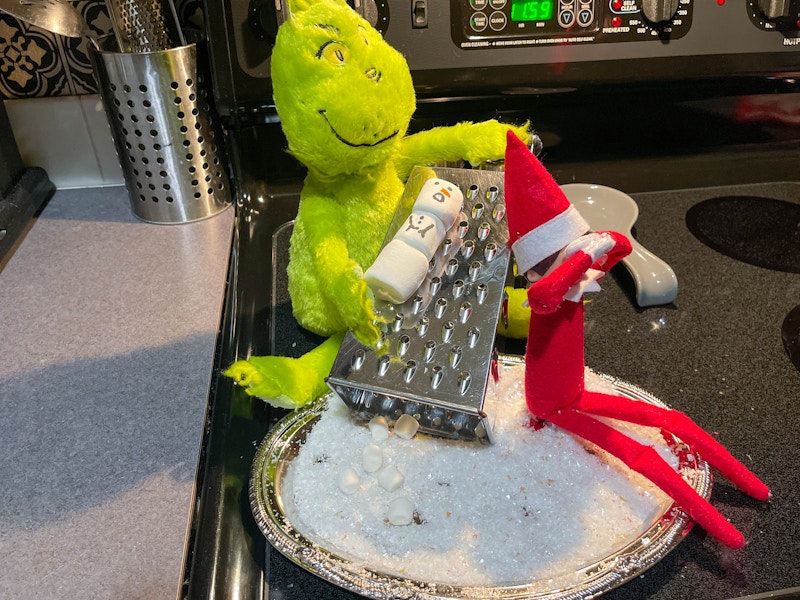 a grinch doll pretending to grate a marshmallow snowman next to a elf on the shelf doll