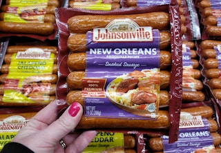 A person's hand holding a package of Johnsonville New Orleans smoked sausages in front of a shelf of more sausages in Kroger.
